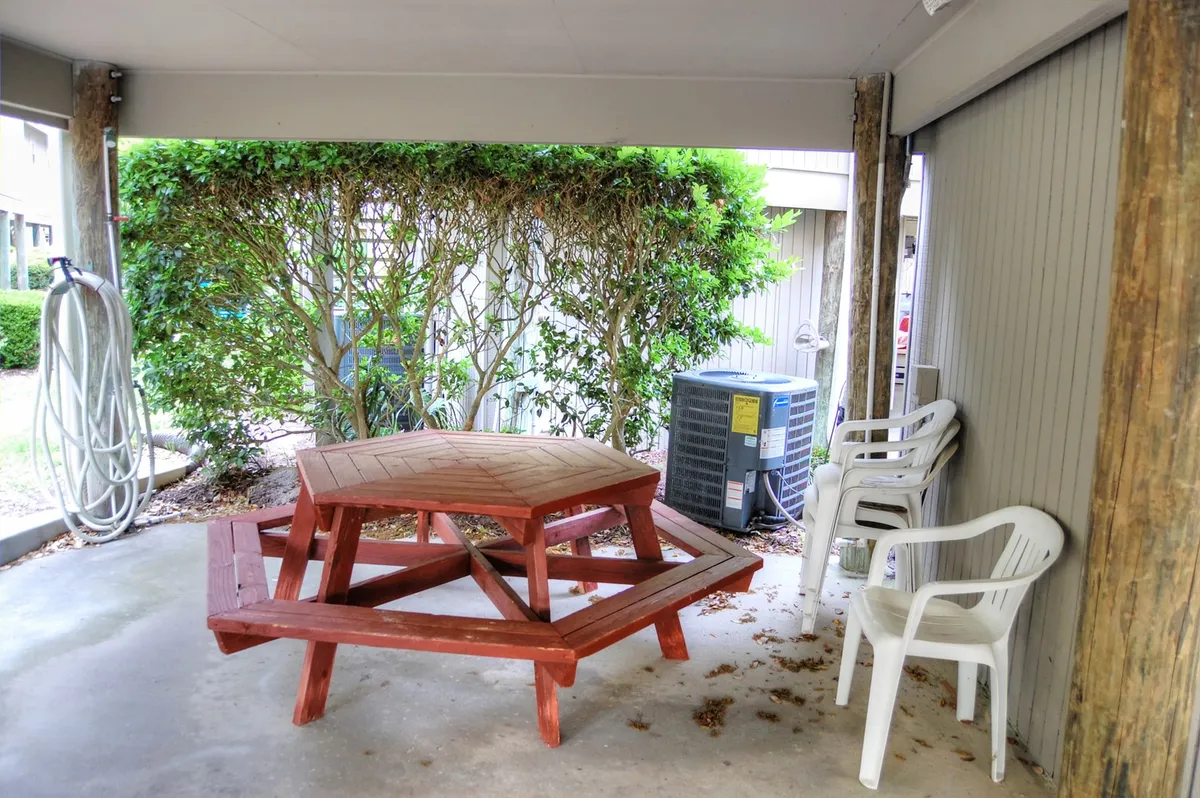 An interior scene featuring a hexagonal picnic table surrounded by white plastic chairs and green hedges visible outside. The relaxed, informal setting evokes the feel of shared meals, socializing, and family gatherings, all in a space that connects to the outdoors.