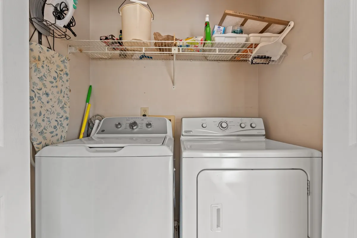A bright, organized laundry area featuring a washing machine and drying machine stacked side by side, with a practical shelf above holding an assortment of cleaning supplies. The space is clearly designed for convenience and everyday efficiency.