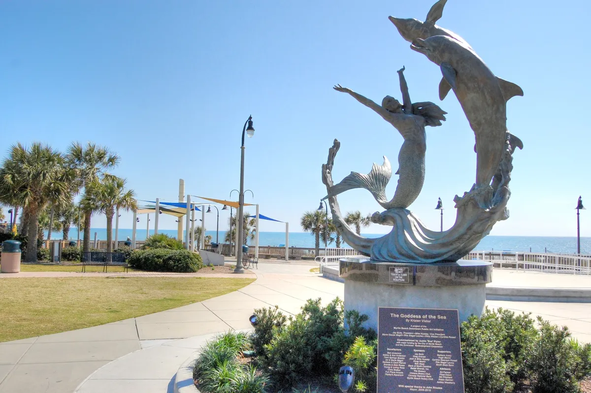 A stunning sculpture of the Goddess of the Sea adorns a beachfront walkway, complemented by playful dolphins and swaying palm trees. The ocean glistens in the background, and visitors can stroll, relax, or take in the view.