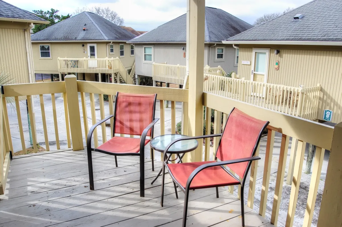 A cozy patio setup with two red chairs and a glass table is nestled among surrounding buildings, offering a private yet inviting space to relax outdoors. The location is calm, ideal for reading or enjoying a sunset.