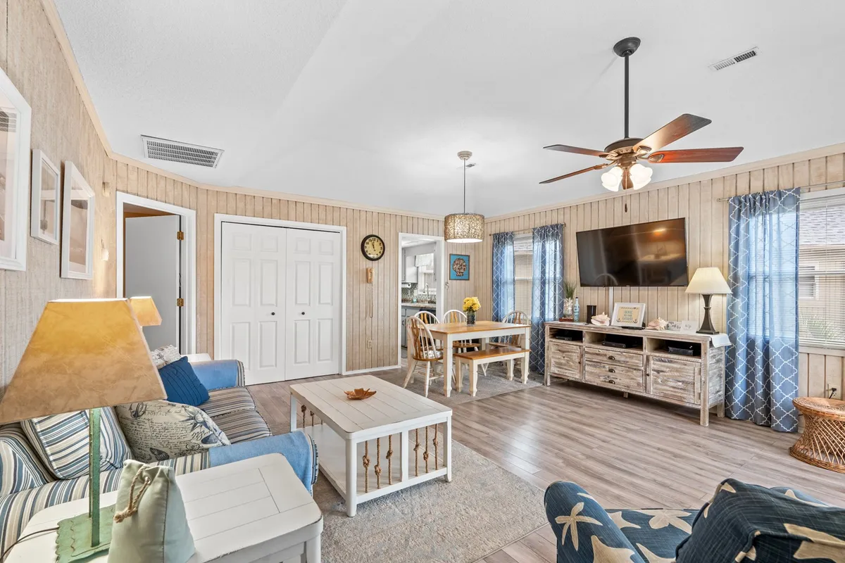 This image shows a welcoming living area featuring a sofa with striped cushions, a wooden coffee table, and a dining area with a round table and chairs. Natural light streams through the windows, highlighting the open floor plan that connects the living and dining spaces. A television on a stand and a decorative lamp complete the cozy scene, perfectly suited for family gatherings and relaxing evenings.