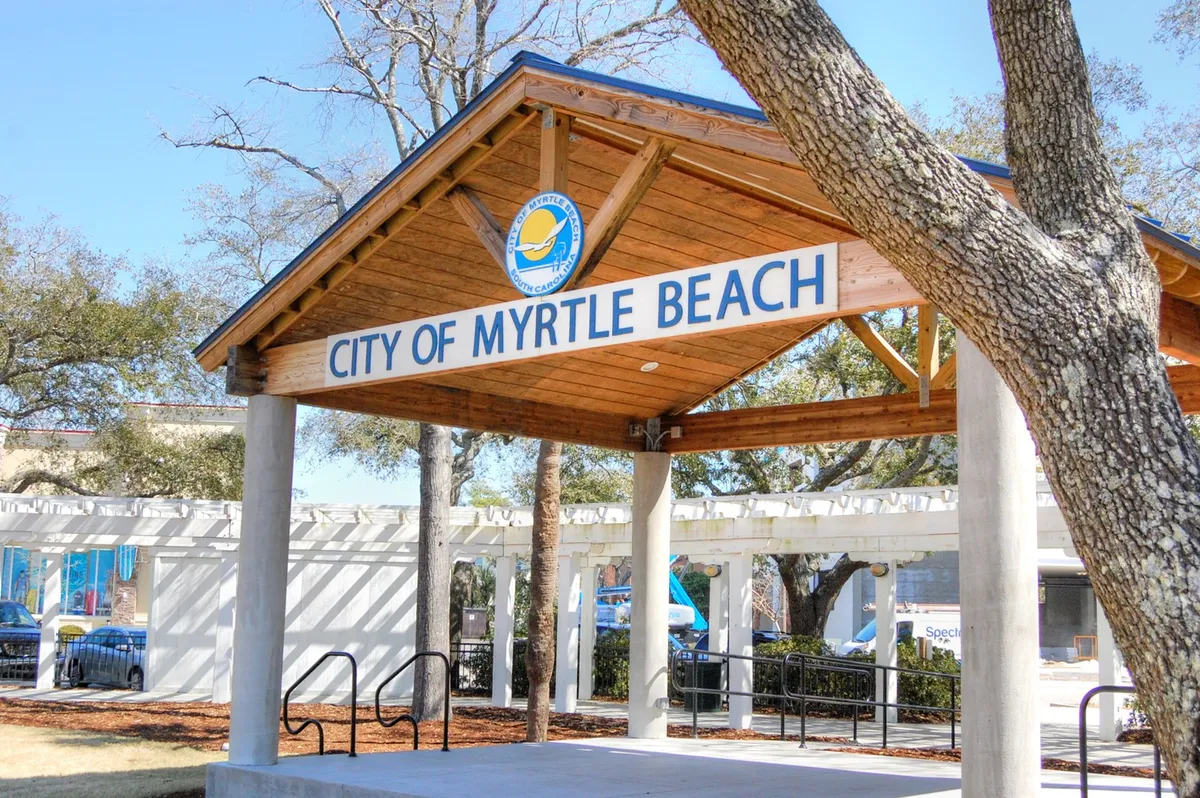 A cheerful exterior scene showing the 'City of Myrtle Beach' pavilion structure under bright skies, surrounded by trees. The setting is clearly oriented toward community gatherings, festivals, and local events, evoking the lively spirit of exploration and group activities nearby.