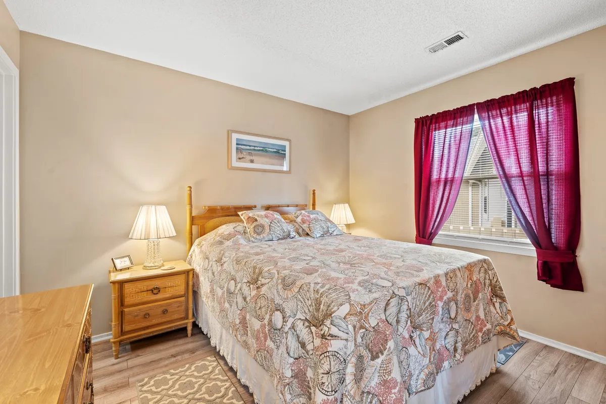 A tranquil bedroom with a bed adorned in shell-patterned bedding, nightstands with warm-lit lamps, framed artwork on the wall, and vibrant red curtains. Sunlight streams through the window, creating a restful and inviting environment.