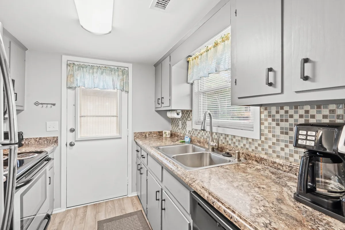 A charming kitchen scene with a mosaic-tiled countertop, kitchen sink, coffee maker, and a sunlit window framed by curtains. The space feels cozy and functional, perfect for morning coffee or prepping holiday meals as a group.