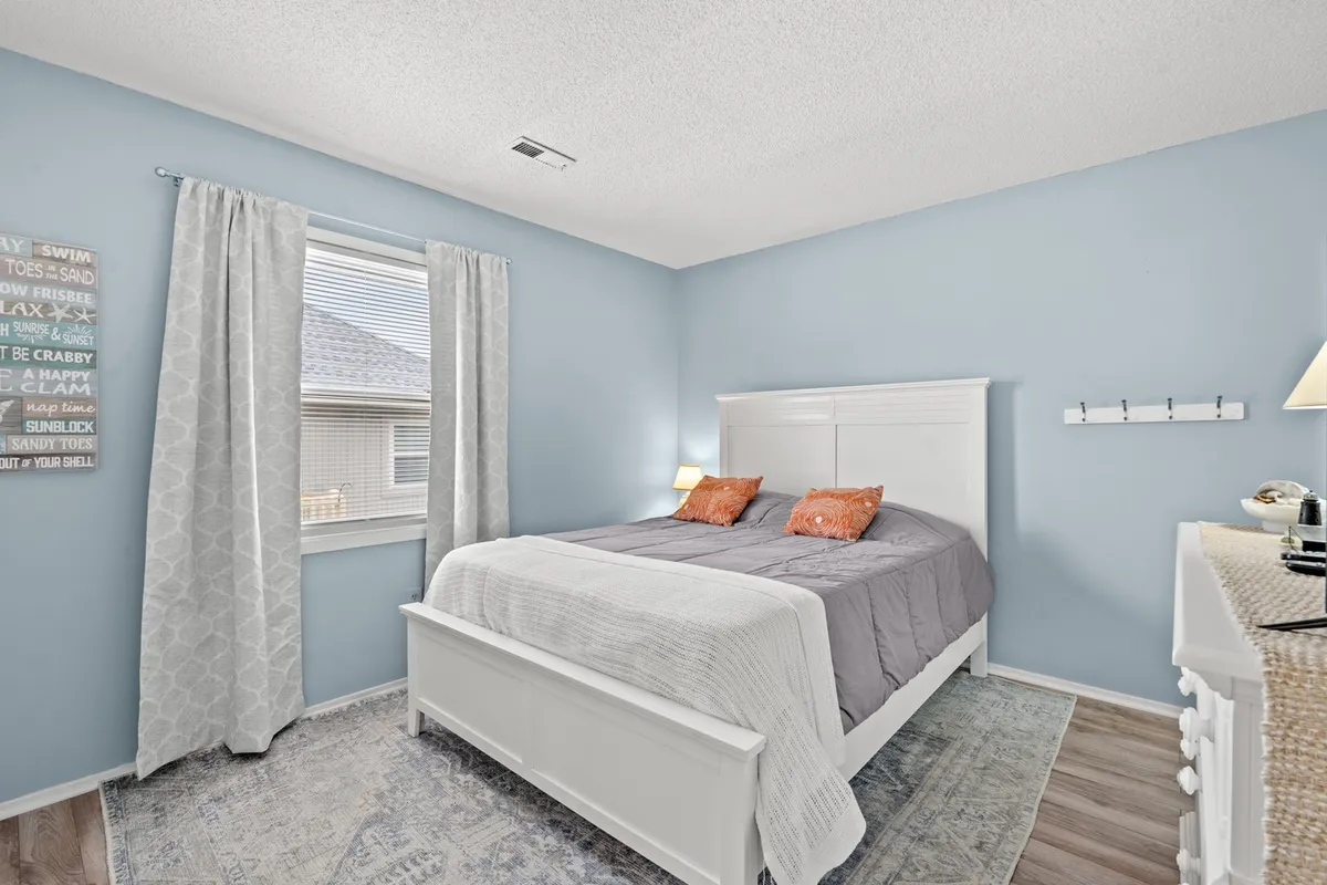 A sunlit bedroom featuring a queen bed, a window allowing natural light to filter in, a decorative wall sign above the bed, and a cozy nightstand. The bedding looks plush and inviting, while the room's overall atmosphere feels serene and calming—perfect for unwinding.