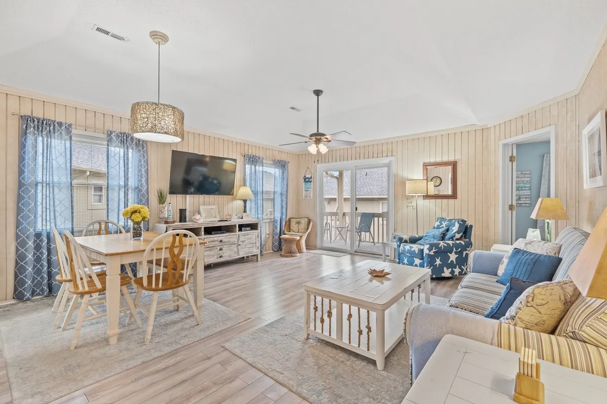 This cheerful, inviting living space features a dining table adjacent to a cozy sofa, with a TV and a door leading to a balcony. The room is filled with light, and the setup encourages socializing, dining, and relaxing. The arrangement highlights both comfort and easy flow between the living and dining areas.