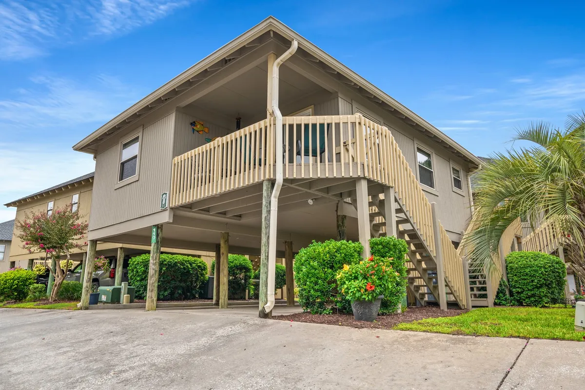 A welcoming exterior view of the stand-alone cottage, with a prominent staircase, lush green bushes, and a balcony perfect for socializing. The scene is vibrant and inviting, suggesting possibilities for relaxing outdoors, sipping coffee, and hosting gatherings with friends or family.