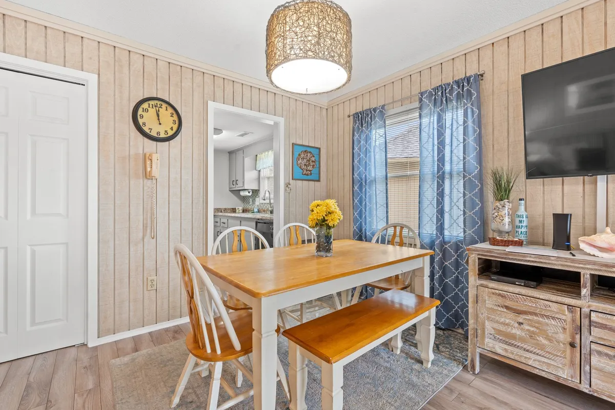 A cozy dining area anchored by a large table beneath a statement chandelier, surrounded by windows and tasteful wall decor. There's a TV nearby, suggesting a space for both meals and casual, laughter-filled gatherings.